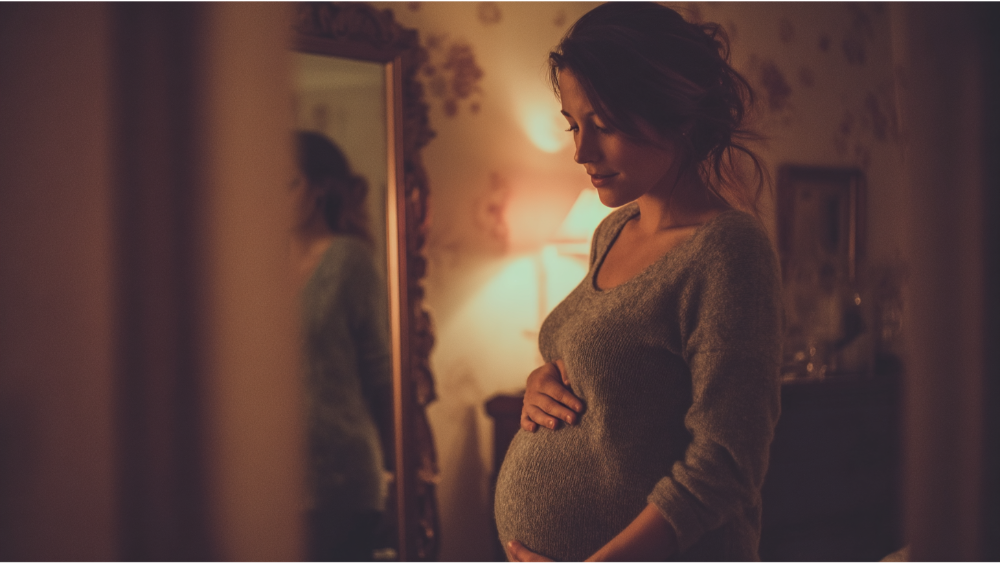 Pregnant woman standing in a softly lit room, touching her belly and gazing down.