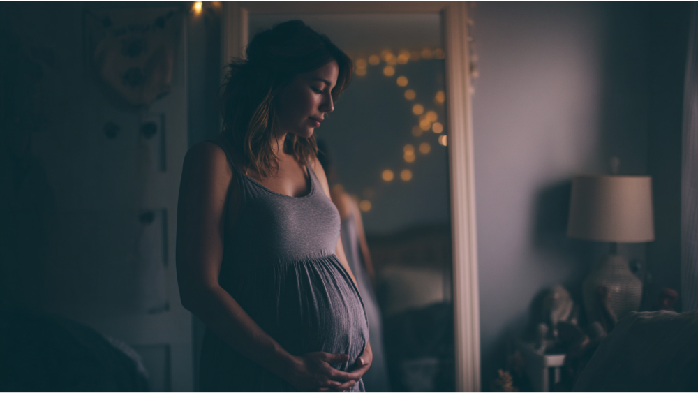 Pregnant woman standing in a dimly lit bedroom, reflected in a mirror with string lights.