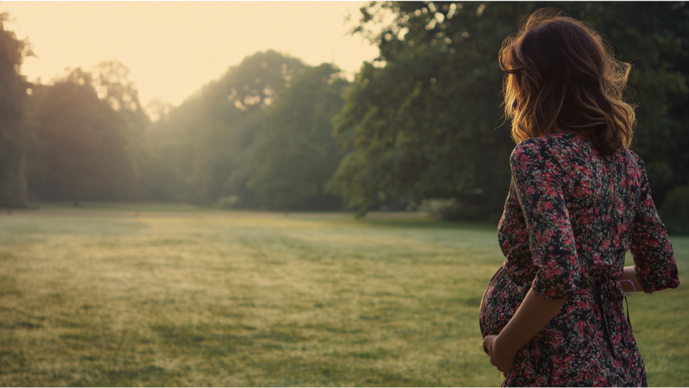 Pregnant woman walking through a sunlit park in a floral dress, holding her belly.