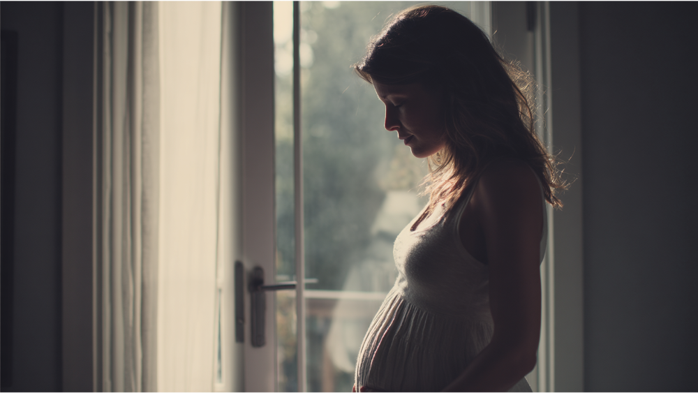 Silhouette of a pregnant woman standing quietly by a window, softly lit from behind.