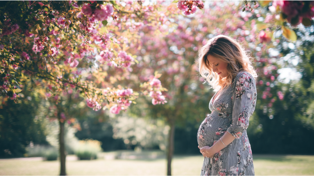 Pregnant woman standing beneath blooming trees in a garden, gently holding her belly.