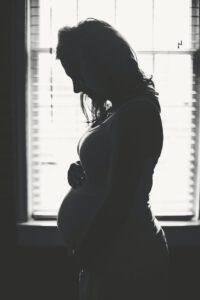 silhouette of pregnant surrogate mother in Brentwood standing indoors next to window, courtesy of Made in the USA Surrogacy agency in California