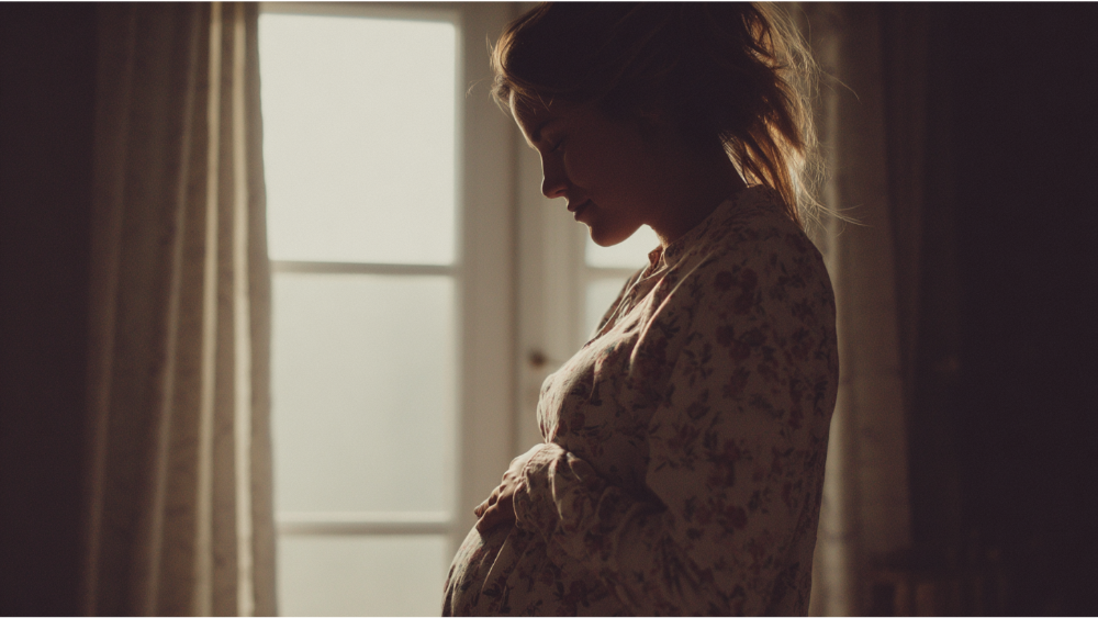 A pregnant woman in a floral top standing near a softly lit window, gently holding her belly.