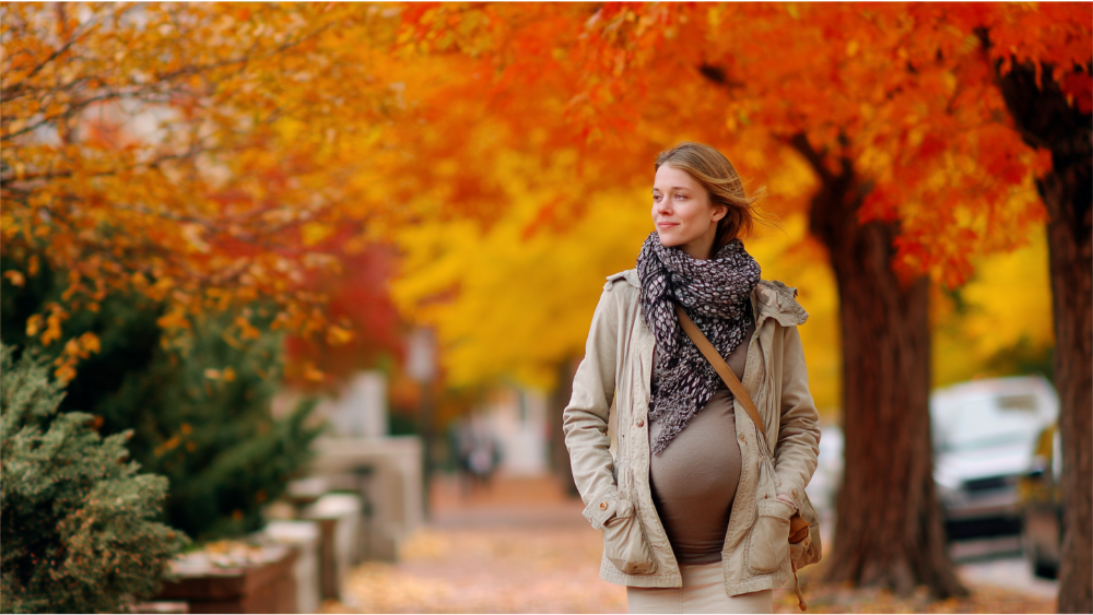 A pregnant woman walking outside on a city sidewalk during autumn, surrounded by vibrant fall foliage.